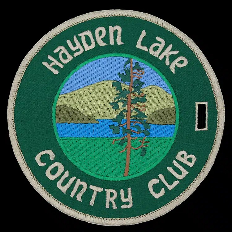 Round embroidered luggage tag with a pine tree scene made for Hayden Lake Country Club, green thread on a light ground.