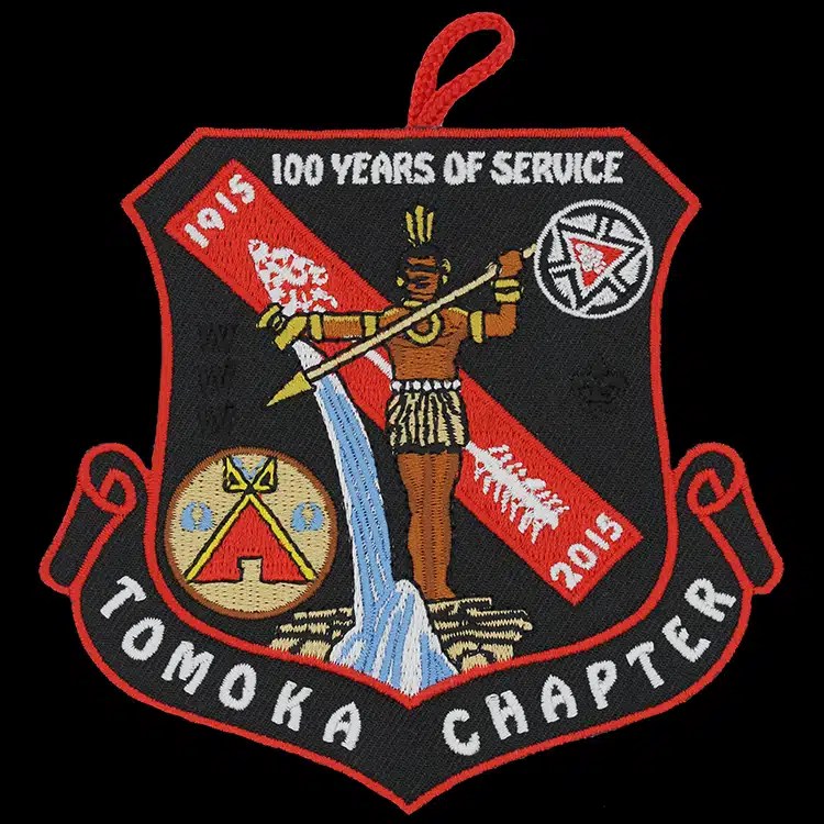Shield-shaped embroidered patch on black twill showing a figure above a waterfall, a red diagonal band with two dates, a red hanging loop at the top and a red merrowed border.
