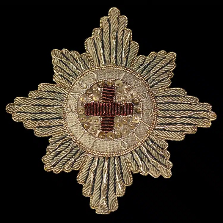 Eight-pointed star badge worked in antique gold bullion wire, with sequins and a red wire cross at the centre, on black.