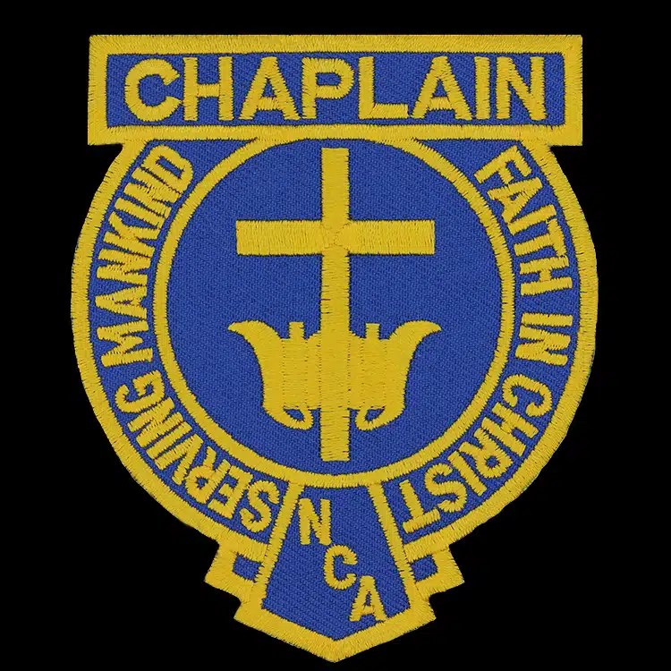 Embroidered shoulder patch in royal blue and gold: a gold cross over a lamp on a blue circle, a title banner across the top and a motto around the ring.