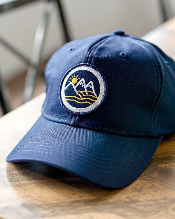 Round embroidered patch with a mountain and sun mark and a merrowed white border on the front of a navy cap.
