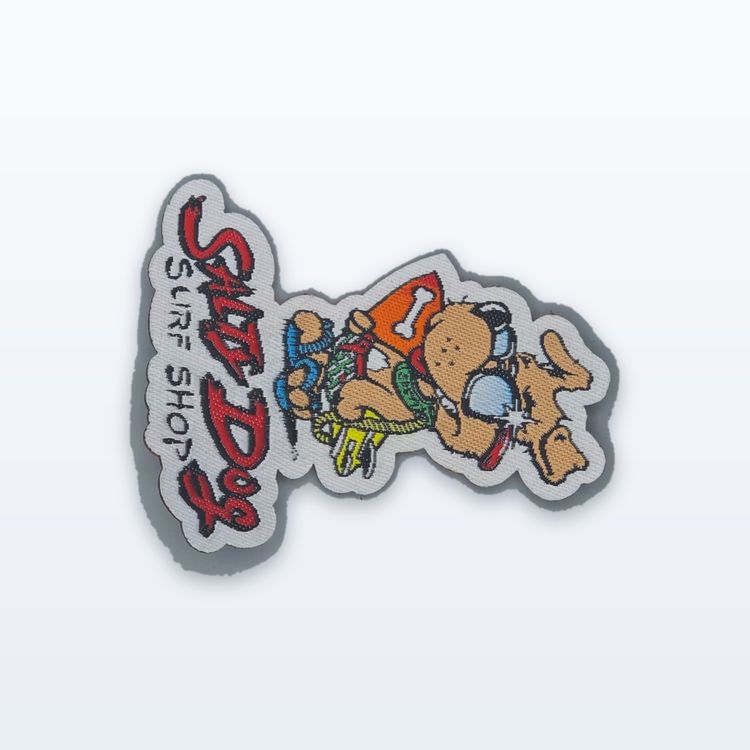 Die-cut woven patch of a cartoon dog in sunglasses and a surf shirt beside a vertical red script wordmark.