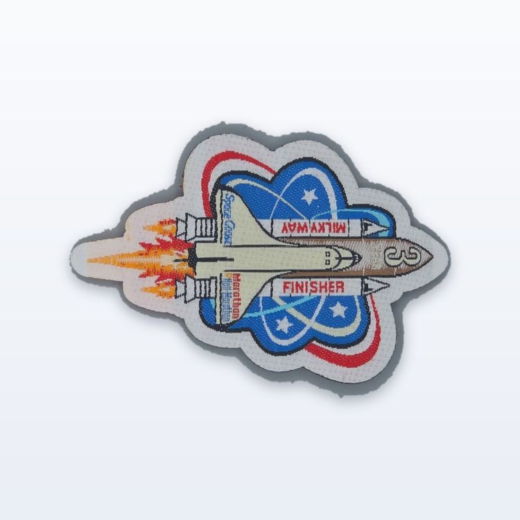 Die-cut woven patch of a space shuttle lifting off over a blue globe with stars, with a small red banner, on a grey woven edge.